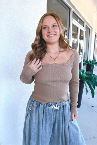A woman wearing a beige ribbed long-sleeve top with lace trim and a light blue denim skirt, standing outdoors in front of a storefront.
