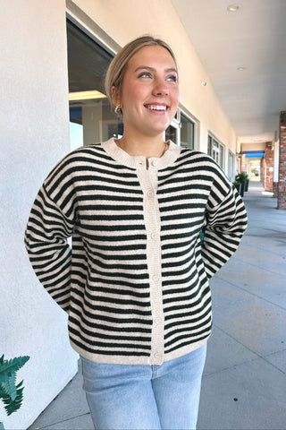 A woman wearing a green and cream horizontally striped knit cardigan with a cream button placket, standing in a covered outdoor walkway.