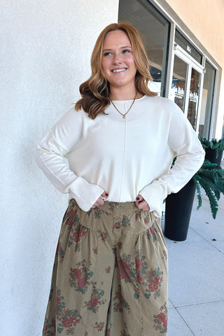 A woman wearing a cream ribbed knit long-sleeve top and high-waisted olive green wide-leg pants with a red and green floral pattern, standing outdoors in front of a building.