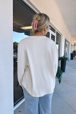 A woman viewed from behind wearing a cream ribbed knit long-sleeve top and light blue jeans, standing on a sidewalk next to a building.