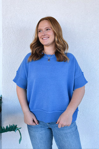 A woman with shoulder-length brown hair stands against a white textured wall wearing a bright blue short-sleeve knit sweater and light blue jeans.