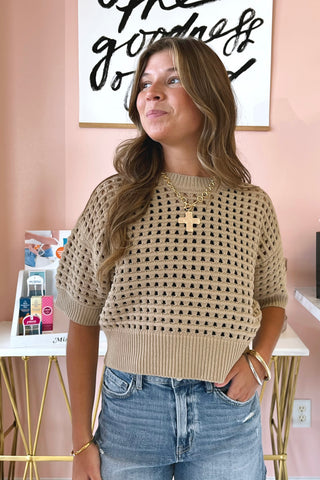 A woman wearing a beige short-sleeve knit sweater with an openwork geometric pattern, paired with blue jeans, standing in a room with a pink wall and a gold-framed sign.