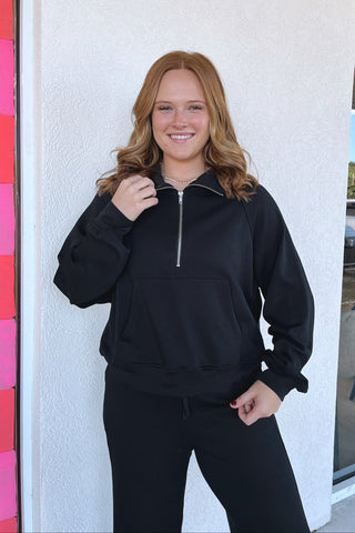 A smiling young woman with long wavy light brown hair stands outdoors wearing a black half-zip pullover hoodie and matching black sweatpants, posing in front of a white stucco wall and a glass storefront window.