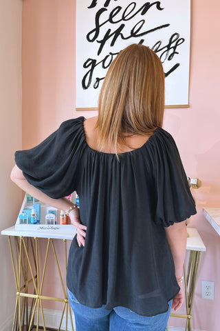 Woman wearing a black off-shoulder top in a room with a pink wall and framed artwork.