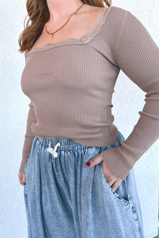 A woman wearing a ribbed long-sleeve scoop-neck top in taupe with a lace trim along the neckline, paired with light blue denim drawstring pants.