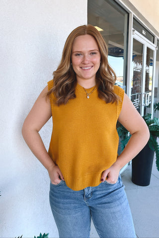 A woman wearing a sleeveless mustard yellow ribbed knit top and light blue jeans, standing outdoors against a white wall next to a glass storefront window.