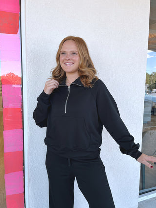 A woman with blonde hair wearing a black half-zip sweatshirt and matching black sweatpants, standing against a white textured wall.