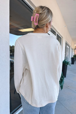 A woman viewed from behind wearing a cream ribbed long-sleeve top and light blue jeans, standing on a covered walkway next to a storefront.