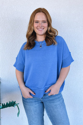 A woman with wavy light brown hair stands smiling, wearing a short-sleeve blue knit sweater with ribbed cuffs and hem, paired with light-wash blue jeans, set against a white textured wall.