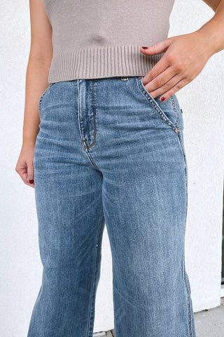 A person wearing light-to-medium wash blue jeans and a beige ribbed knit top, standing against a white textured wall.
