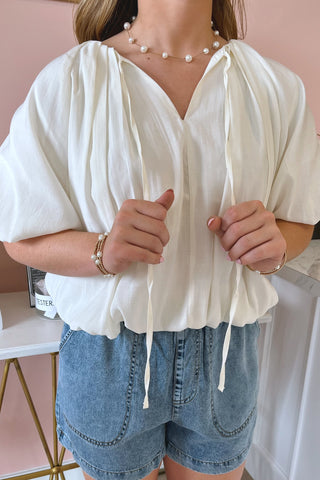 Ella-Morgan is wearing an oversized, ivory, ruched bubble top and soft washed chambray shorts. It has large balloon sleeves and drapes softly. She is wearing a pearl necklace and matching pearl bracelets.