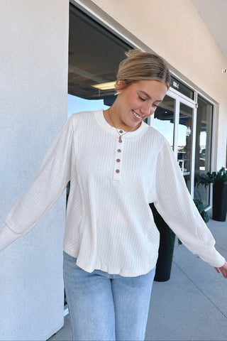 A woman wearing a white ribbed long-sleeve henley top with brown buttons and light blue jeans, standing outdoors in front of a storefront.