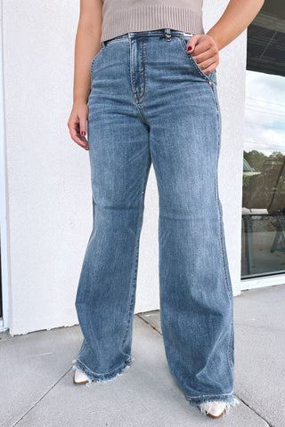 A person wearing light-wash blue wide-leg denim jeans with a frayed hem, paired with a beige ribbed top and white pointed shoes, standing outdoors in front of a white wall and glass window.