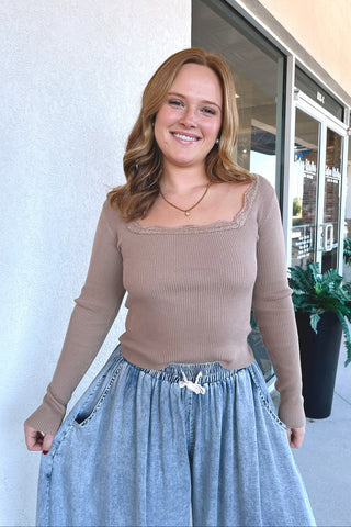 A woman wearing a beige ribbed long-sleeve scoop-neck top with lace trim and a light blue denim skirt, standing outdoors in front of a storefront.