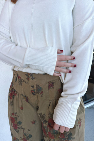 A person wearing a cream ribbed knit sweater with wide ribbed cuffs and hem, paired with tan floral pants, shown from the chest down.