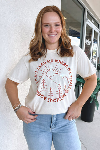 A smiling woman with red hair stands outdoors wearing a cream-colored graphic t-shirt with a red circular design featuring trees, a sun, and the text 'SPIRIT LEAD ME WHERE MY TRUST IS WITHOUT BORDERS'. She is also wearing light blue jeans.