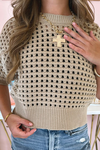 A woman wearing a beige open-knit short-sleeve sweater with a gold cross pendant necklace, paired with blue jeans.