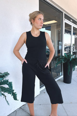A woman wearing a black sleeveless crop top and matching black wide-leg cropped pants stands outdoors against a white wall next to a glass storefront entrance.