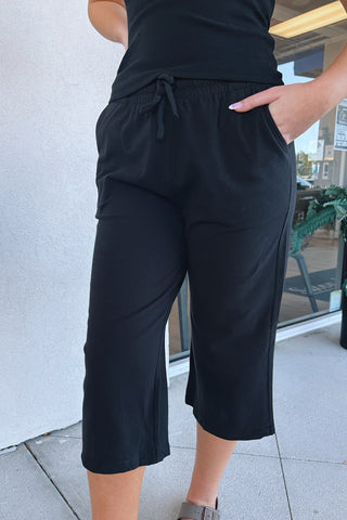 A person wearing black cropped drawstring pants with side pockets, paired with a black top and gray sandals, standing on a tiled walkway near a storefront.
