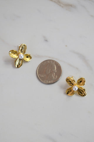 Gold flower-shaped earrings with a pearl center on a marble surface next to a penny for scale.