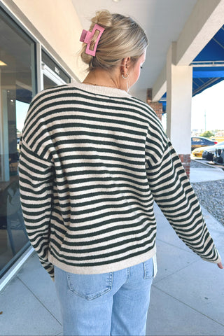 A woman viewed from behind wearing a green and cream horizontally striped knit sweater and light blue jeans, standing on a covered outdoor walkway.