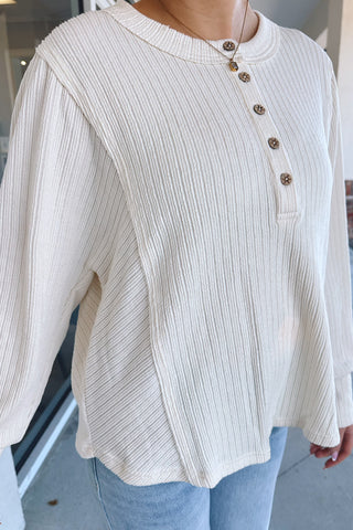 A person wearing a cream ribbed knit long-sleeve henley top with brown buttons, paired with light blue jeans, standing outdoors near a building entrance.