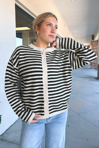 A woman wearing a green and cream striped cardigan with a cream button placket, paired with light blue jeans, standing in a covered outdoor walkway.