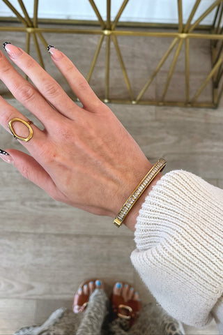 Hand wearing a gold ring and bracelet on a wooden floor.