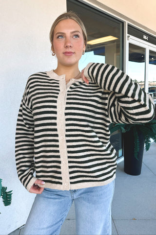 A woman wearing a green and cream striped knit cardigan with a buttonless placket, paired with light blue jeans, standing outdoors in front of a building entrance.