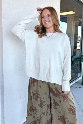 A woman stands outdoors in front of a building, wearing a cream-colored long-sleeve knit top and wide-leg beige pants with a red and green floral pattern.