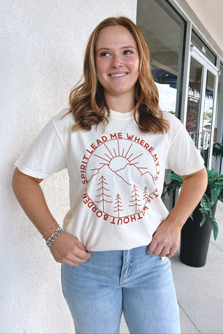 A woman wearing a cream-colored graphic t-shirt with a red circular design that reads 'SPIRIT LEAD ME WHERE MY TRUST IS WITHOUT BORDERS' around a sun and pine tree illustration, paired with light blue jeans, standing outdoors against a white wall.