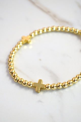 Gold beaded bracelet with a cross charm on a white marble surface