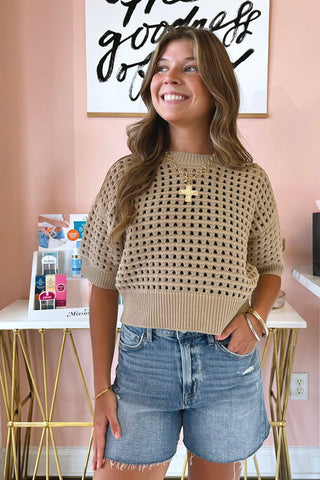 A woman wearing a beige short-sleeve knit sweater with a star-patterned open weave and ribbed hem, paired with denim shorts, standing in a room with a pink wall and a gold-framed art print.