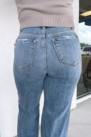 Rear view of a person wearing light-wash blue jeans with a beige ribbed sweater, standing outdoors near a storefront window.