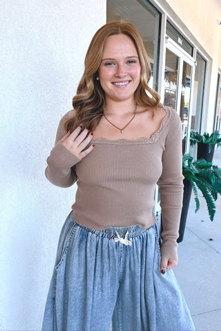 A woman smiling while wearing a beige ribbed long-sleeve top with a scoop neckline and lace trim, paired with a light blue denim skirt, standing outdoors in front of a building.