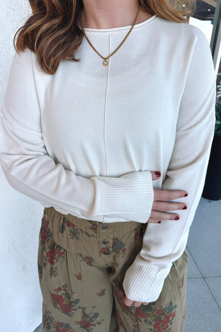 A woman wearing a cream-colored long-sleeve knit top with ribbed cuffs and a center seam, paired with olive green floral pants.