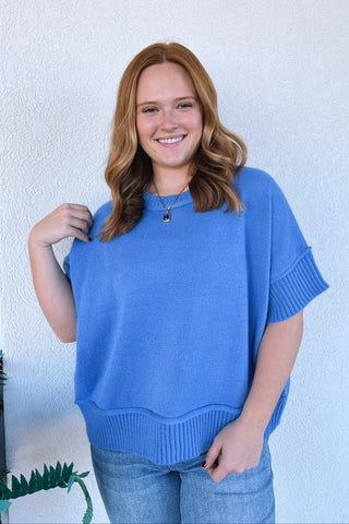 A smiling woman with red hair stands against a white textured wall wearing a bright blue short-sleeve knit sweater and light blue jeans.