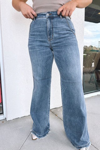 A person wearing high-waisted, wide-leg blue denim jeans with a frayed hem, standing outdoors on a sidewalk next to a white wall and storefront window.