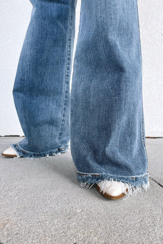 Close-up of a person wearing wide-leg blue denim jeans with frayed hems, paired with white pointed-toe flats, standing on a concrete surface against a light wall.