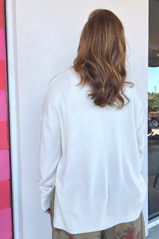 A woman viewed from behind wearing a white knit sweater with a ribbed hem, standing against a white wall.