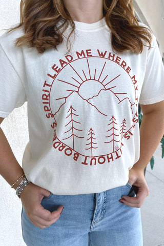 A person wearing a white graphic T-shirt with a red circular mountain, sun, and pine tree design and the text 'SPIRIT LEAD ME WHERE TRUST IS WITHOUT BORDERS' visible around the print. The photo is cropped from the neck down, showing the shirt and light blue jeans against an outdoor wall background.